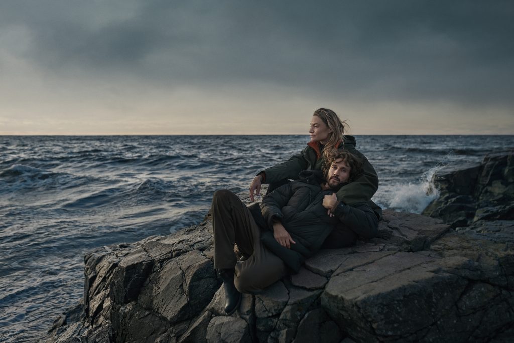 Photo of man and woman sat on rock by the sea wearing Didriksons coats